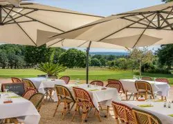 Terrasse avec des chaises en osier, des tables nappées, des parasols blancs et un jardin arboré.
