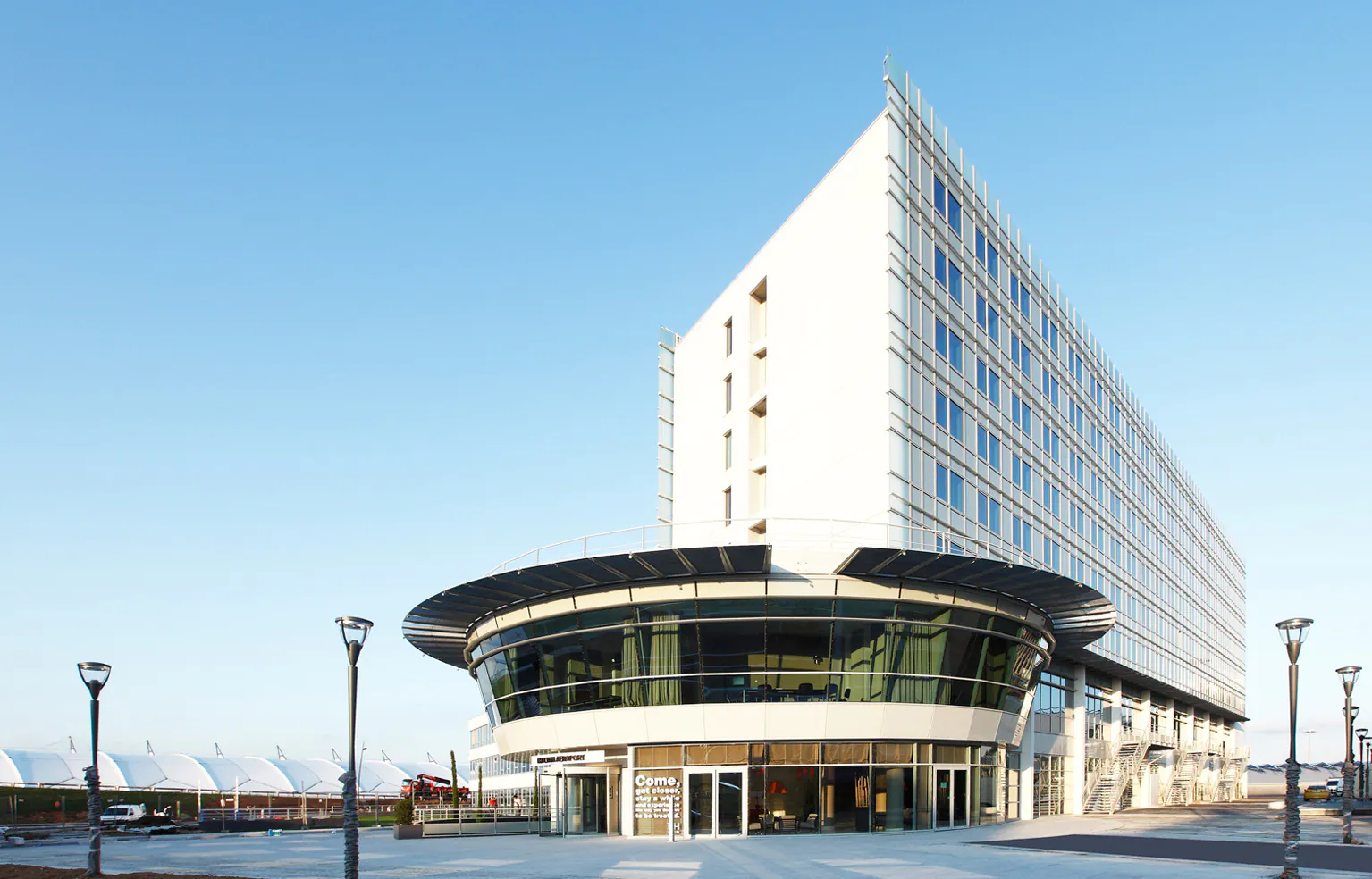 en:: a six-story white rectangular building with blue-framed windows, featuring a large two-story curved section on the left, seminar rooms upstairs and a lounge on the ground floor fr:: immeuble rectangulaire blanc de six étages avec fenêtres bleues, doté à son extrémité gauche d’un large arrondi sur deux niveaux, avec salles de séminaire à l’étage et salon lounge au rez-de-chaussée