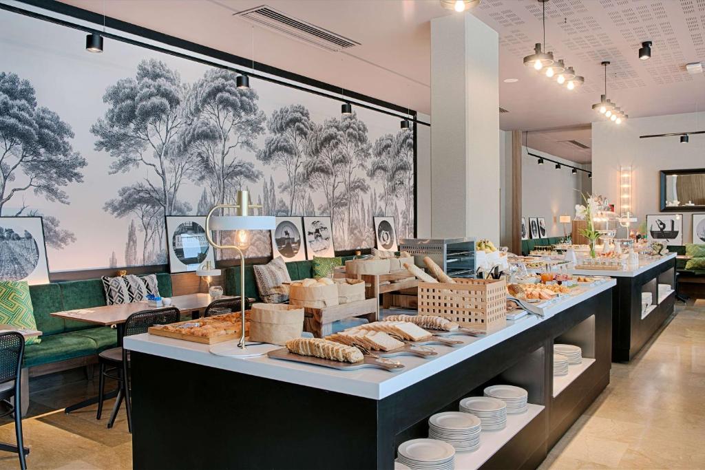 en:: breakfast buffet with bread and pastries in the foreground, and a dining area with green banquette seating, cushions, square tables, and wooden chairs in the background fr:: buffet de petit-déjeuner avec espace pains et viennoiseries au premier plan, et en arrière-plan banquette aux assises vertes avec coussins, tables carrées et chaises en bois