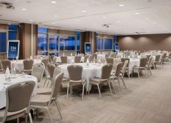 en:: reception room with round tables dressed in white, beige chairs, an NH Hotels sign, and blue evening reflections in the floor-to-ceiling windows fr:: salle de réception avec tables rondes nappées de blanc, chaises beiges, affiche NH Hotels et reflets bleus de la nuit dans les baies vitrées