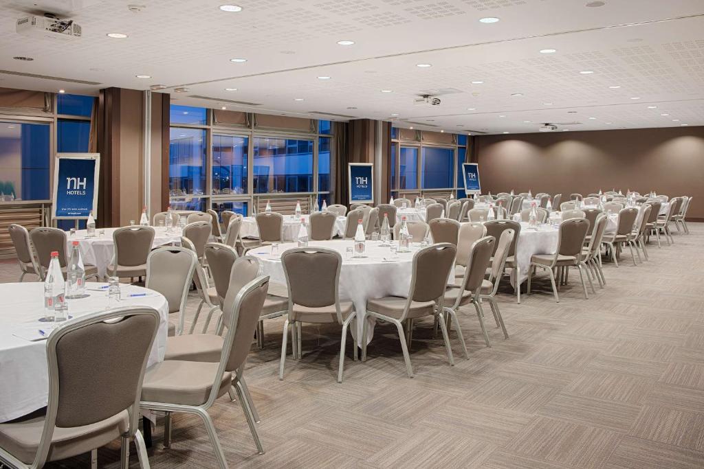 en:: reception room with round tables dressed in white, beige chairs, an NH Hotels sign, and blue evening reflections in the floor-to-ceiling windows fr:: salle de réception avec tables rondes nappées de blanc, chaises beiges, affiche NH Hotels et reflets bleus de la nuit dans les baies vitrées
