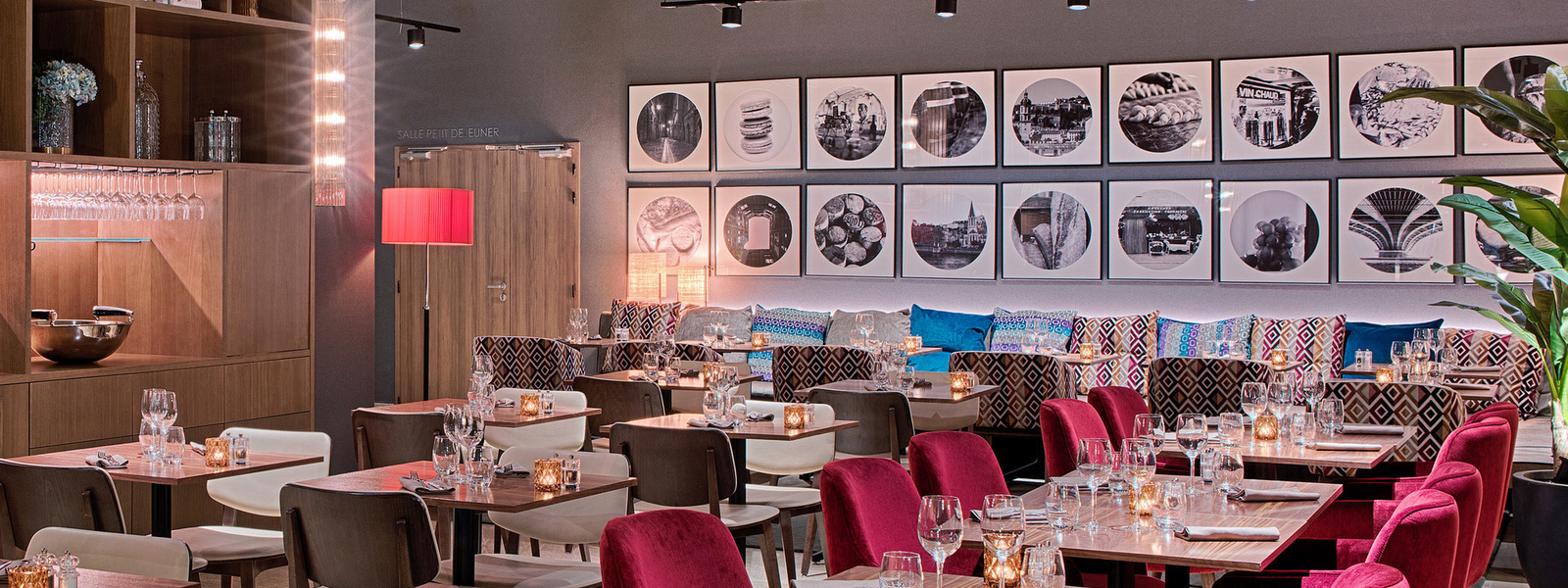 en:: a seminar hotel restaurant with a wall-side banquette, colorful cushions, framed photographs, raspberry-colored chairs, and square tables fr:: salle de restaurant d’un hôtel séminaire avec banquette le long du mur, coussins multicolores, photographies encadrées, chaises couleur framboise et tables carrées
