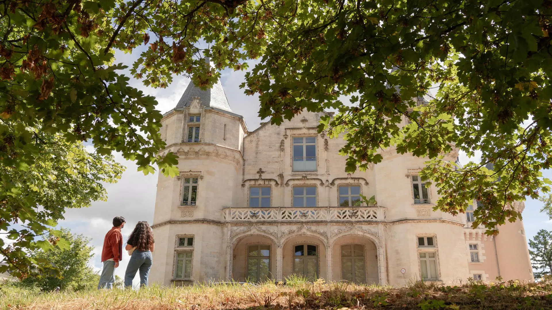 Château médiéval entouré d’une végétation luxuriante, avec deux personnes vues de dos en train de l’observer, dans une ambiance estivale.