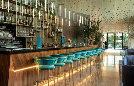 A modern bar with high chairs upholstered in electric turquoise blue fabric and gold legs. A black marble and wood countertop, with pendant lights. In the back, a gold-colored display unit for beverages, cocktails, and glasses, set against a black-and-white lacquered floor.
