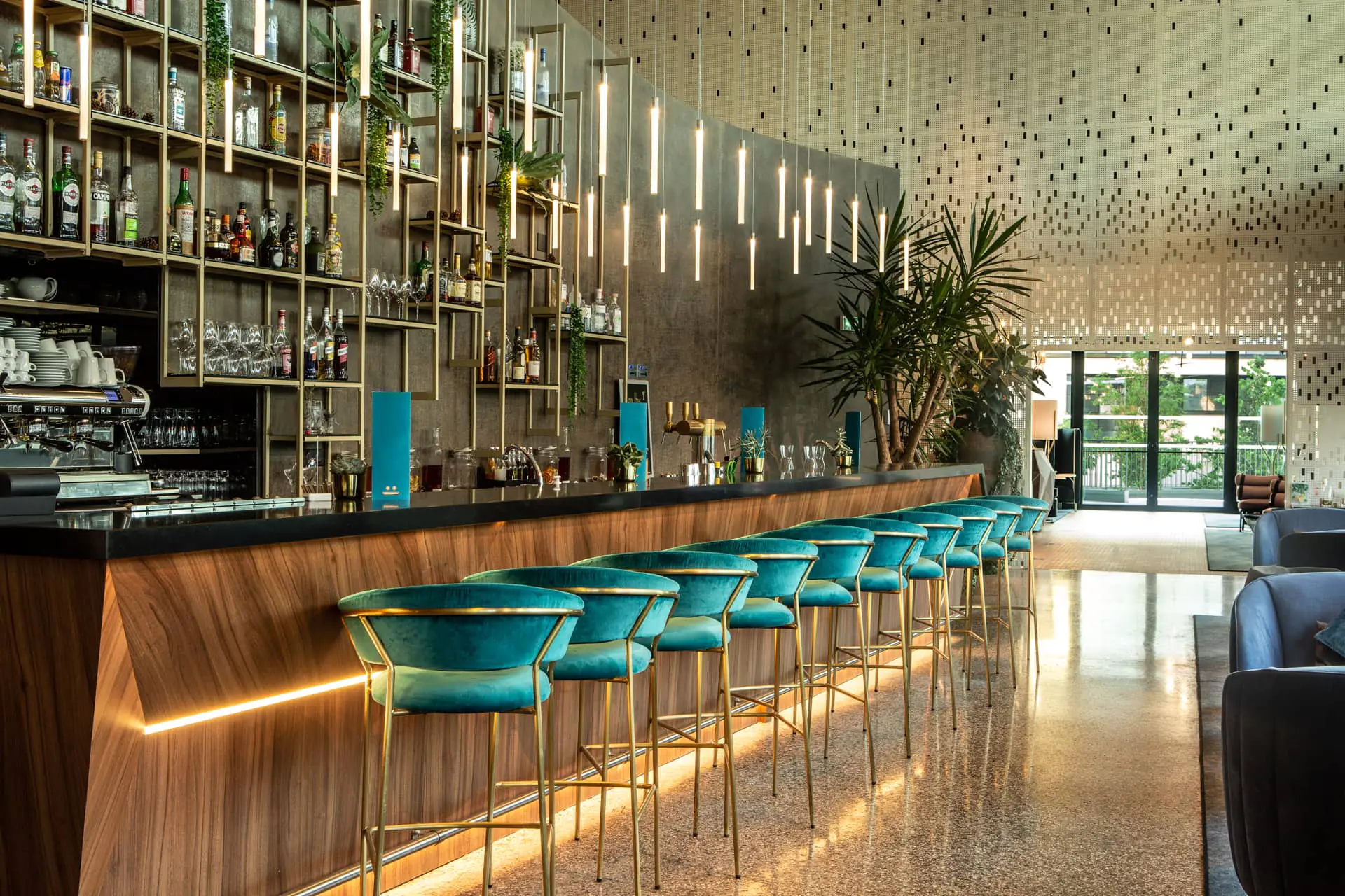 A modern bar with high chairs upholstered in electric turquoise blue fabric and gold legs. A black marble and wood countertop, with pendant lights. In the back, a gold-colored display unit for beverages, cocktails, and glasses, set against a black-and-white lacquered floor.