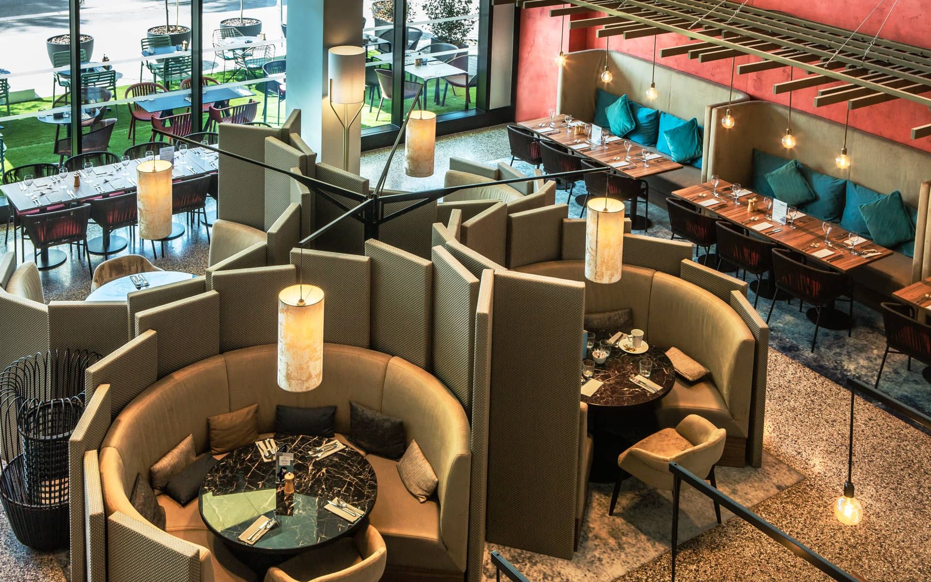 A view of the restaurant from above, showing circular, partitioned dining areas, sofas along the walls, and a view of the outdoor terrace. Pendant lights hang above the sofas, and there are white and gray rugs and carpeting.
