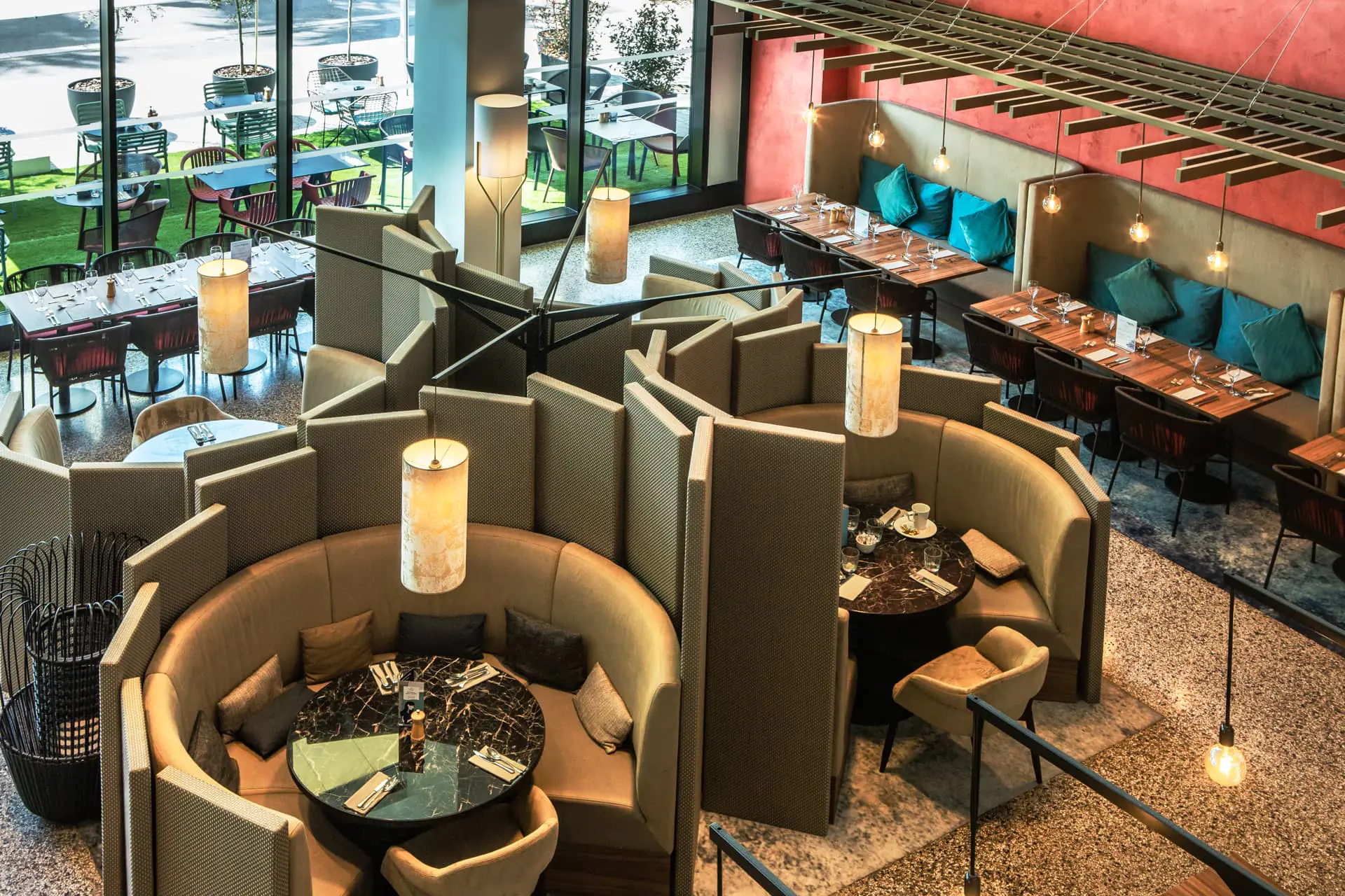 A view of the restaurant from above, showing circular, partitioned dining areas, sofas along the walls, and a view of the outdoor terrace. Pendant lights hang above the sofas, and there are white and gray rugs and carpeting.