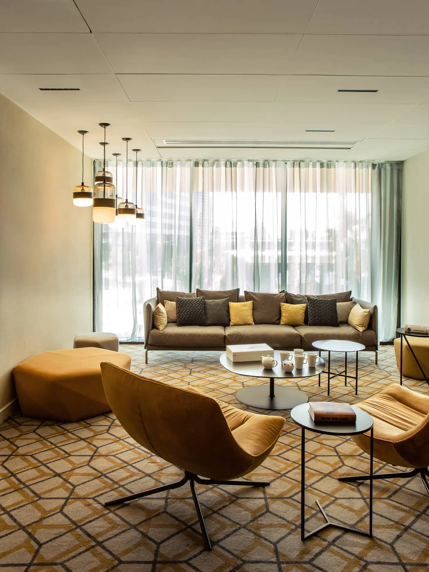 A lounge area with a large sofa and fabric armchairs, sea-green sheers, and cream-colored walls. Coffee tables, tea, ottomans, and modern pendant lights. Carpet with a geometric pattern in yellow, black, and cream.