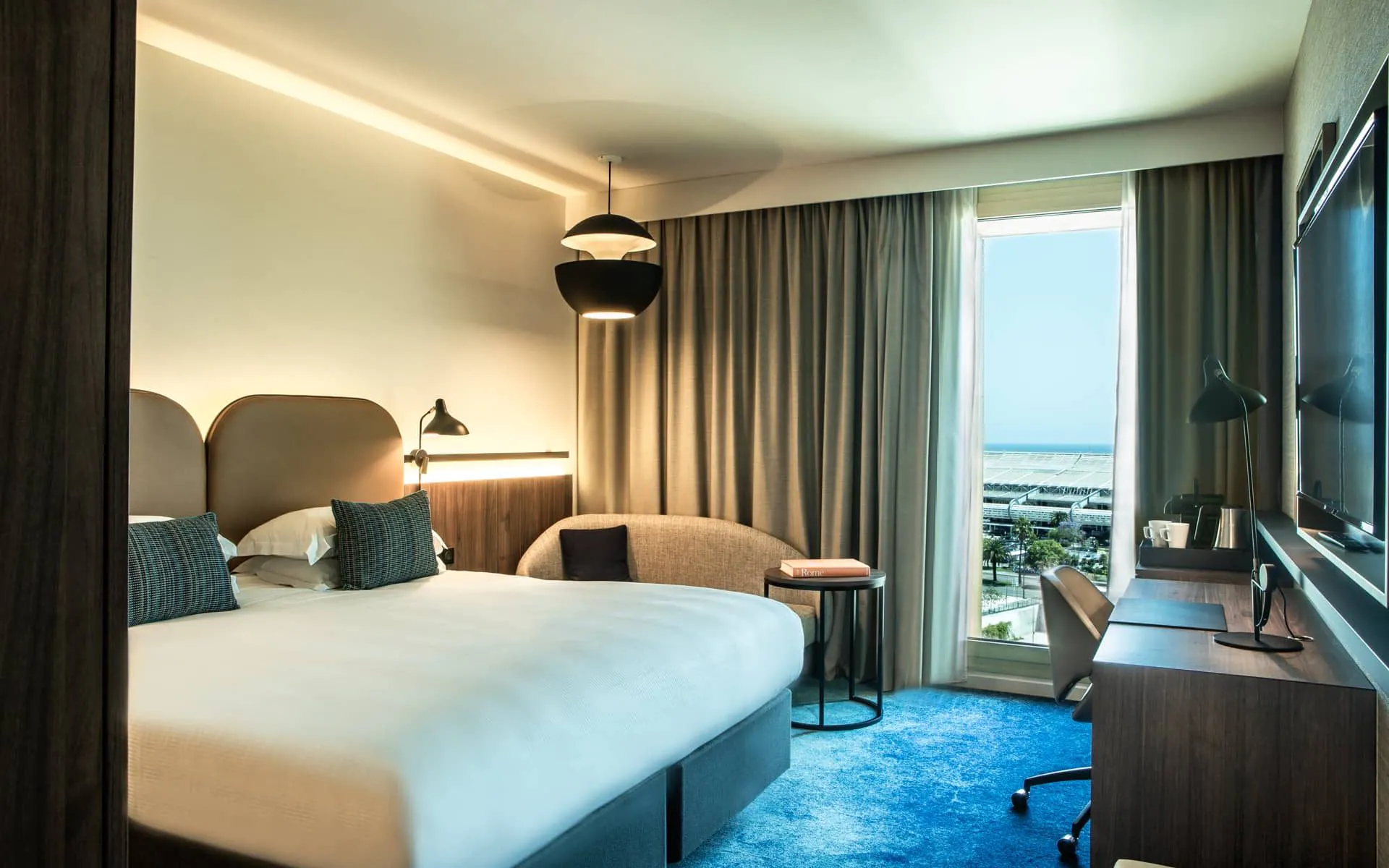 Bedroom with a double bed and two taupe-gray leather headboards, cream-colored walls, and electric sky-blue carpet. View of the sea and the city of Nice, with a desk area and a wall-mounted TV facing the bed. Taupe-gray blackout curtains, a sofa and table area, and modern lighting fixtures.