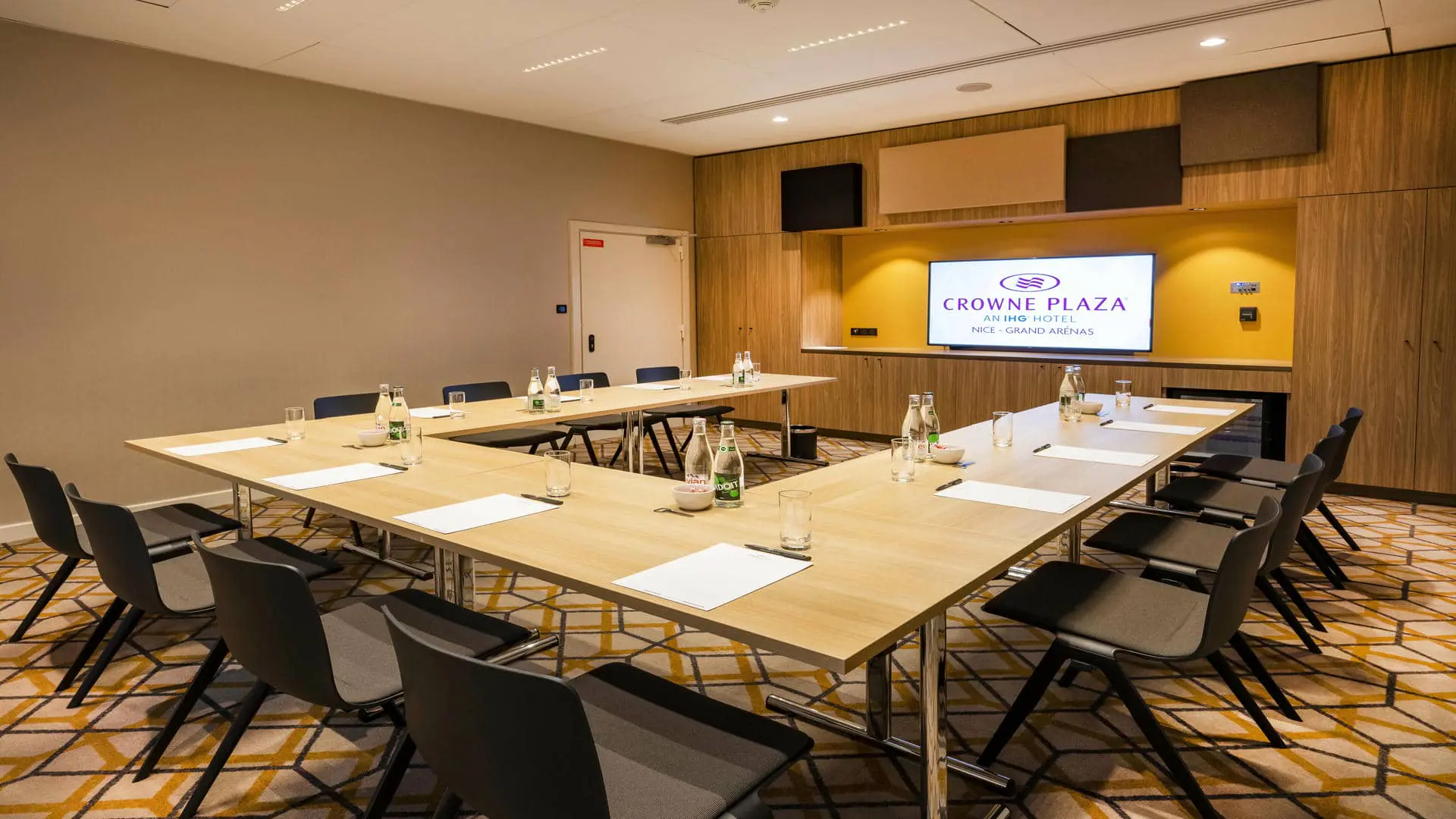 Meeting room with wooden tables arranged in a U-shape and black chairs. Water, notepads, and pens are available; a projection screen is surrounded by wooden cabinets. The walls are cream and gray, and the carpet features a geometric pattern in black, yellow, and cream.