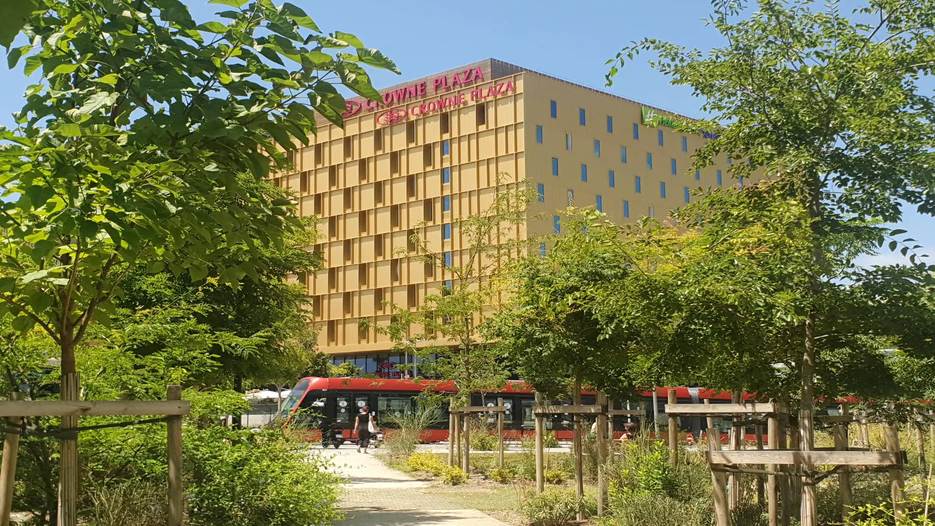 The imposing yellow facades of the Crowne Plaza Nice and the Holiday Inn Express. A tram passes in front of the hotels, surrounded by greenery and parks.