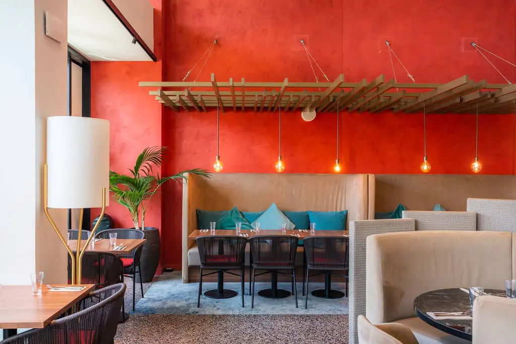 A restaurant with red walls, large leather sofas, and tables and chairs. Blue-and-white rugs, black-and-white carpet, and modern cream-colored light fixtures. Wooden structures above the dining areas, with pendant lights.