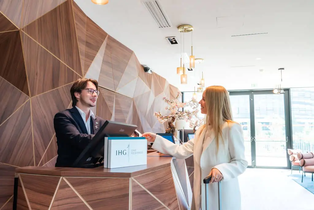 A wooden hotel reception desk with geometric relief. A receptionist hands a key to a woman in a white business suit, accompanied by her suitcase, in front of the IHG sign. Warm lighting and colors.