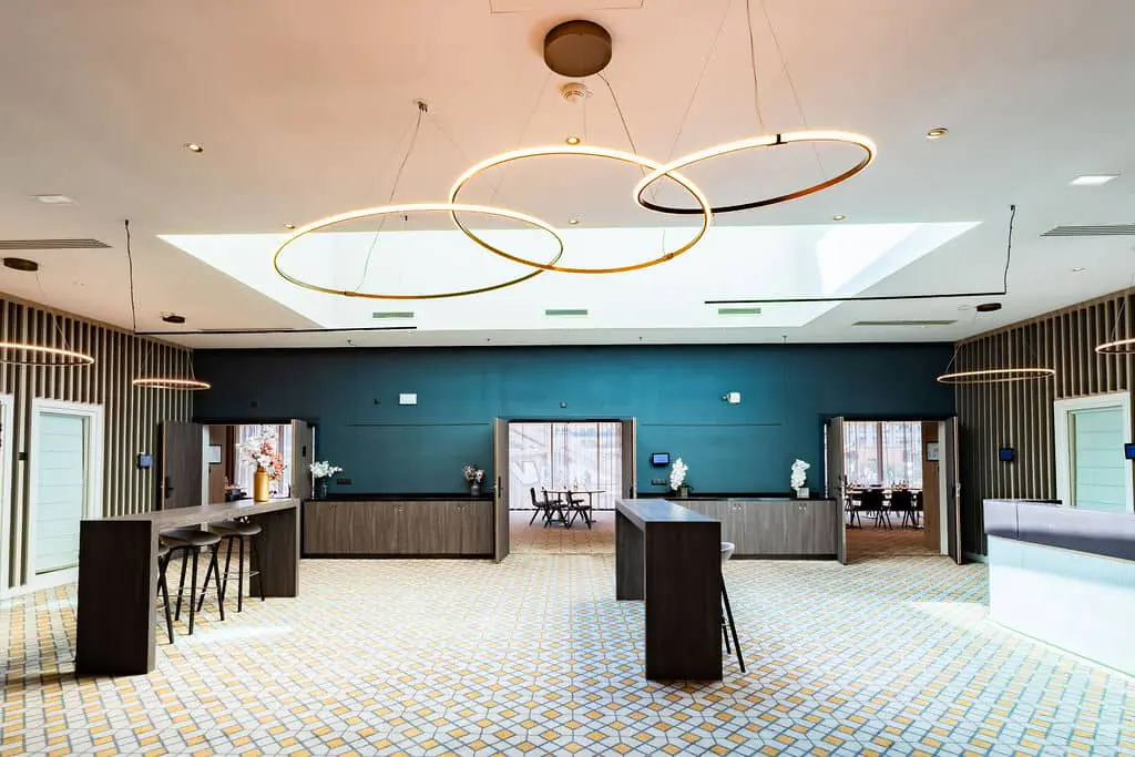 A waiting area serving three meeting rooms, with two sideboards on either side of the doors. Two tables and bar stools, modern lighting fixtures, a teal wall, and a black wall decorated with wooden sticks. Carpet with a geometric pattern in black, yellow, and cream.