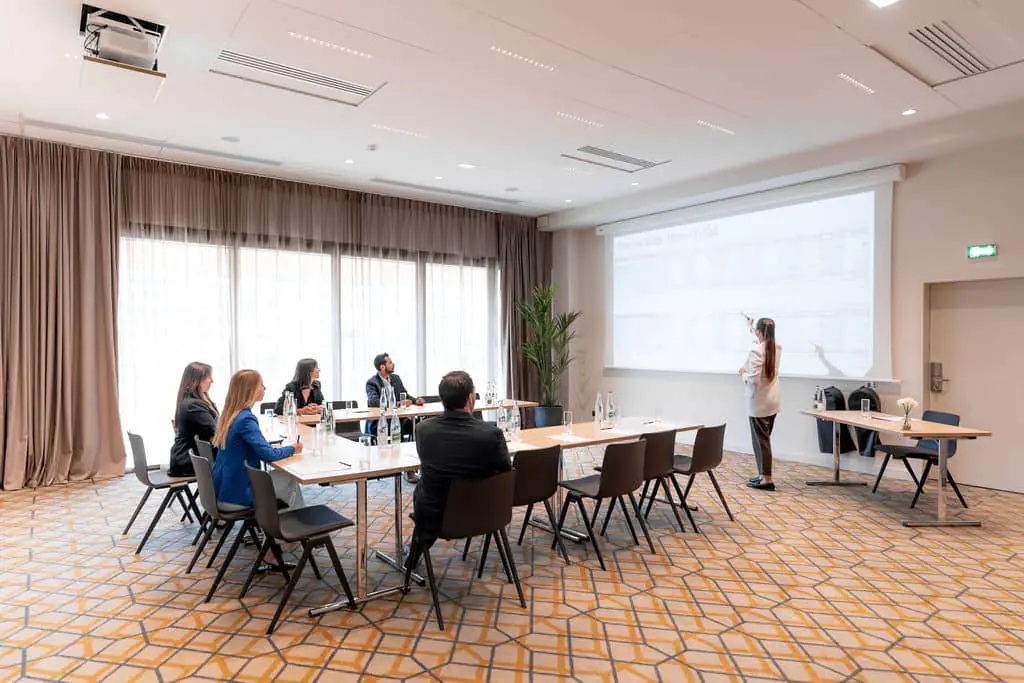 Meeting room with a projector, tables, and chairs. Water, notepads, and pens are available. Cream-colored blackout curtains, cream-colored walls, and carpet with a black-and-yellow geometric pattern. Emergency exit.