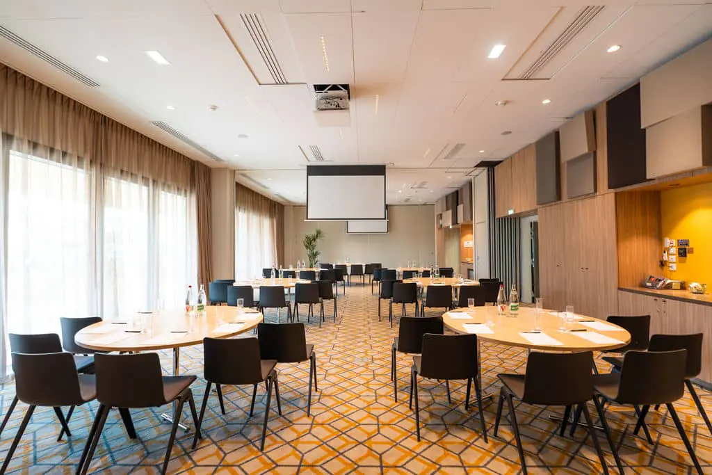 Large meeting room with round tables and black chairs. Bottled water, notepads, pens, a kettle, and tea are available. Two projectors, cream-colored blackout curtains, cream-colored walls, and carpet with a geometric pattern in black, yellow, and cream.