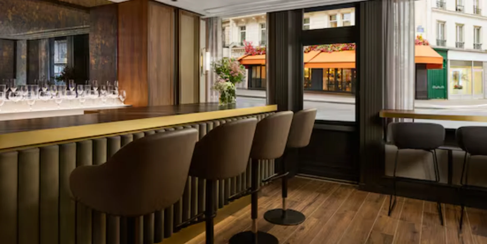 Élégant bar d’hôtel de luxe à l’atmosphère intimiste, donnant sur une charmante rue parisienne, avec un long comptoir raffiné aux finitions en laiton brossé et aux détails en bois cannelé sombre, accompagné de fauteuils hauts aux tons taupe. Les riches boiseries en bois sombre, l’éclairage chaleureux et la belle présentation de la verrerie créent une ambiance sophistiquée et accueillante. De grandes baies vitrées baignent l’espace de lumière naturelle et offrent une vue sur l’animation de la rue, tandis que de délicates compositions florales apportent une touche fraîche et raffinée.