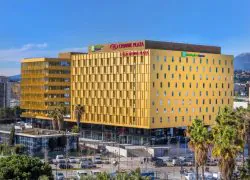 A distant view of the Crowne Plaza Nice, with a yellow facade consisting of two imposing buildings. Surrounding it are a parking lot, the tram, and palm trees.