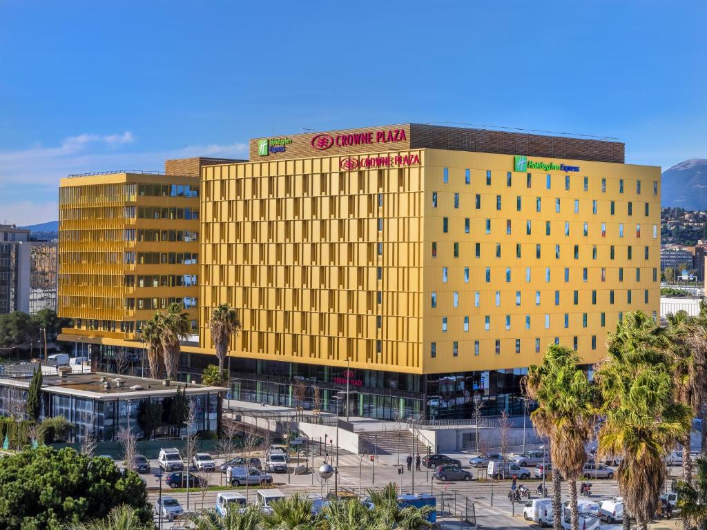 A distant view of the Crowne Plaza Nice, with a yellow facade consisting of two imposing buildings. Surrounding it are a parking lot, the tram, and palm trees.