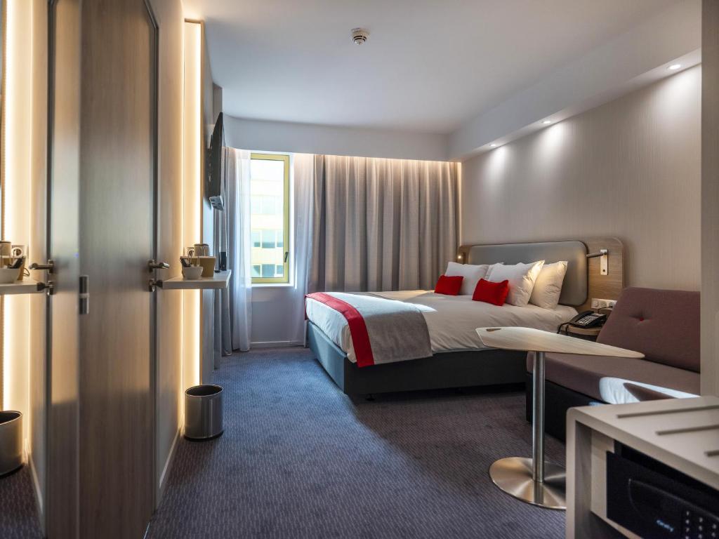 Room with two separate beds and a wooden headboard upholstered in gray leather. Wall-mounted TV, electric kettle, tea, sofa, and safe provided. Cream-colored blackout curtains and walls, taupe carpet. Connecting door to another room.