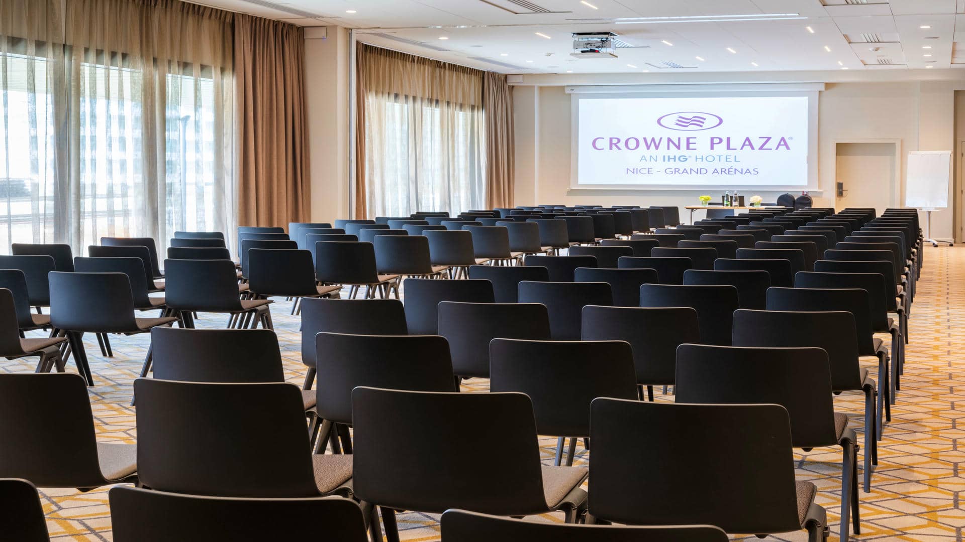 A large meeting room with rows of black chairs, a projector, and whiteboards. Brown-and-cream sheers and blackout curtains, cream-colored walls, and carpet with a geometric pattern in yellow, black, and cream.