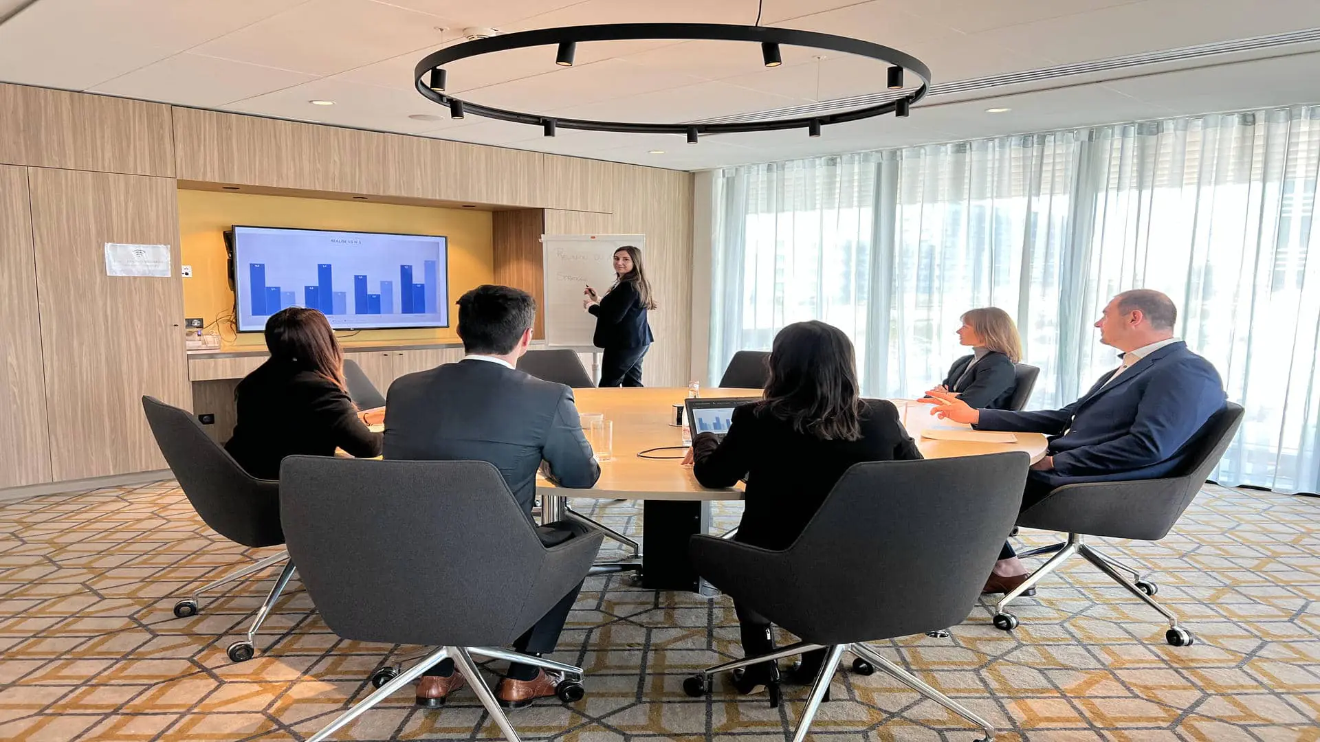 A meeting room with a round table and black fabric armchairs on casters, occupied by people in a meeting. Aqua-blue sheer curtains, a large television, whiteboards, and walls with a wood-grain and cream finish. Carpet with black, cream, and yellow geometric patterns