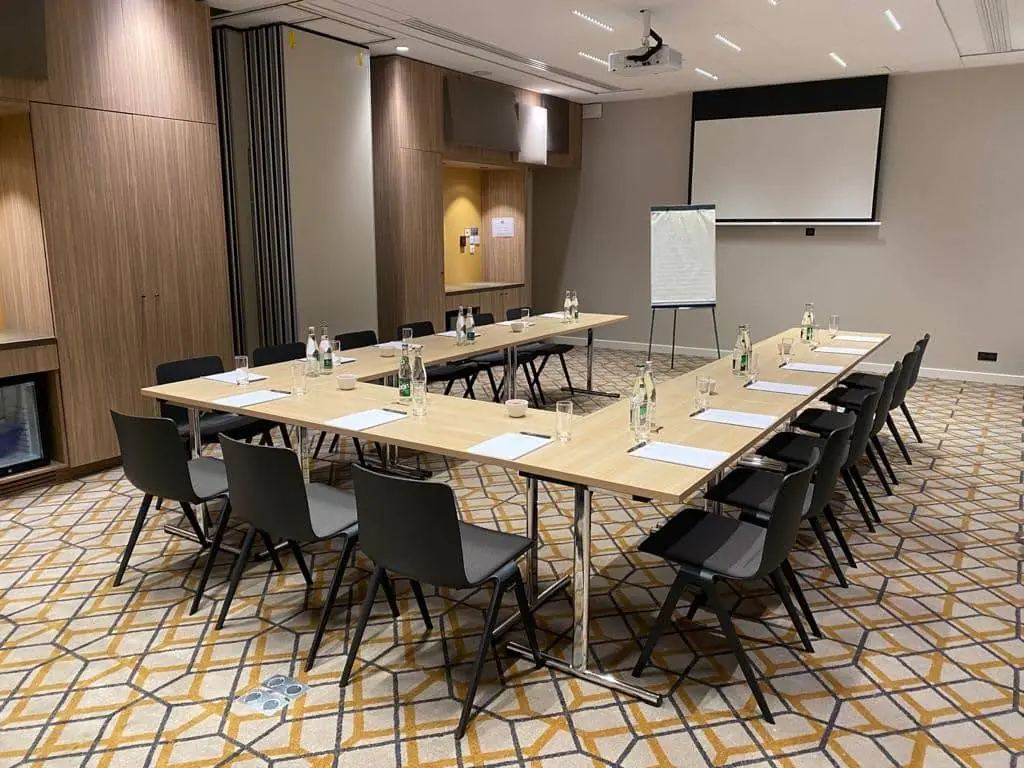 Meeting room with three tables arranged in a U-shape, black chairs, notepads, pens, glasses, and bottles of water. Whiteboard and projector; carpet with black, yellow, and cream geometric patterns. Movable wall on one side.