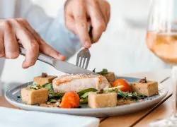 Gros plan sur une expérience gastronomique raffinée, avec un plat soigneusement présenté dans une grande assiette blanche. Celui-ci se compose d’un morceau de poisson doré à la cuisson, accompagné de tomates cerises rôties, de jeunes pousses vertes et de garnitures dorées disposées avec soin, le tout agrémenté de fines herbes et de sauce. Un convive découpe le poisson à l’aide d’un couteau et d’une fourchette, tandis qu’un verre de vin rosé est posé sur la table en bois, à côté d’une serviette blanche soigneusement pliée. La lumière douce et la présentation soignée du plat créent une atmosphère élégante et accueillante.