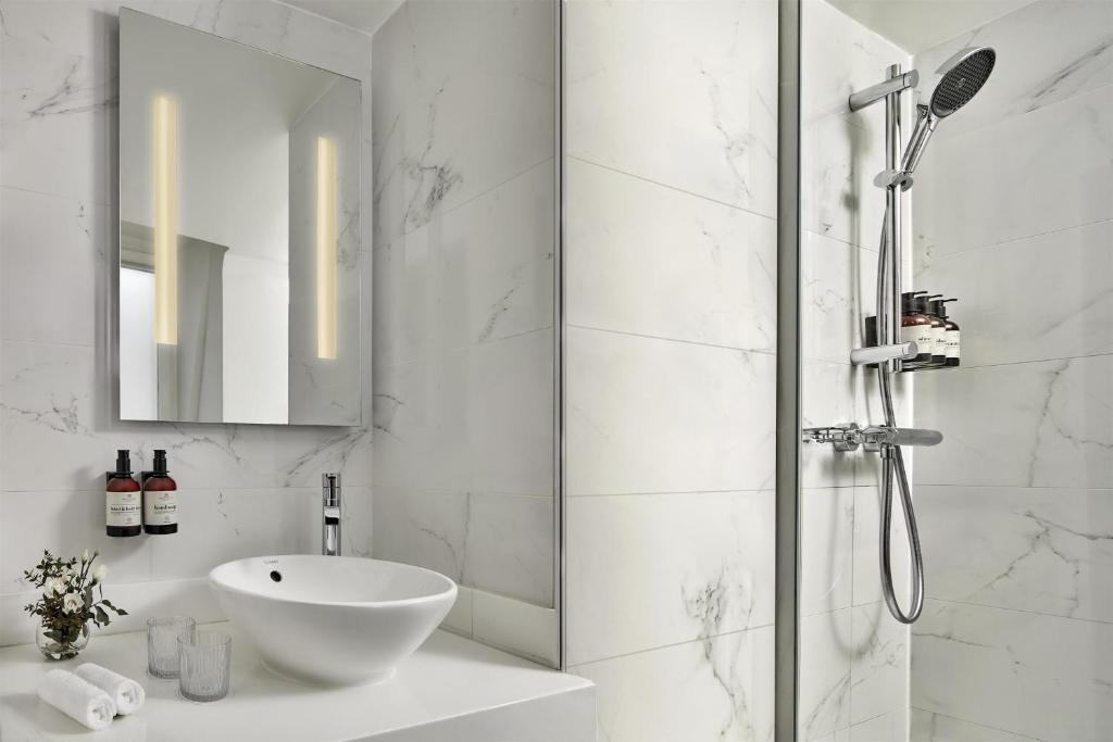 Bright, modern bathroom featuring a large rectangular backlit mirror, a white vessel sink set on a spacious light-colored vanity, and elegant accessories. The walls are covered with white marble-effect tiles with delicate grey veining. A walk-in shower with a glass partition and chrome shower column completes the space, with several bottles arranged on the holder. Rolled white towels and a small floral arrangement add a warm and refined touch.