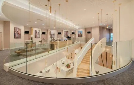 Intérieur d’hôtel comprenant une vaste mezzanine contemporaine surplombant le hall principal. L’étage supérieur est bordé de garde-corps incurvés en verre avec de fines structures métalliques et aménagé avec de petites tables, des chaises et des assises basses de salon. Plusieurs photographies architecturales encadrées décorent les murs beige clair, accompagnées d’écrans noirs fixés au mur. De nombreuses suspensions fines aux finitions dorées descendent du haut plafond blanc et confèrent à l’ensemble une atmosphère élégante et raffinée. À droite, un escalier incurvé aux marches en bois clair et aux parois latérales blanches relie les deux niveaux, avec un éclairage linéaire intégré. Le sol est recouvert d’une moquette grise, tandis que le niveau inférieur présente un revêtement clair, du mobilier moderne et des éléments décoratifs. L’association du verre, du bois clair, des tons neutres et de l’éclairage chaleureux donne à l’espace une apparence lumineuse, sophistiquée et aérée.