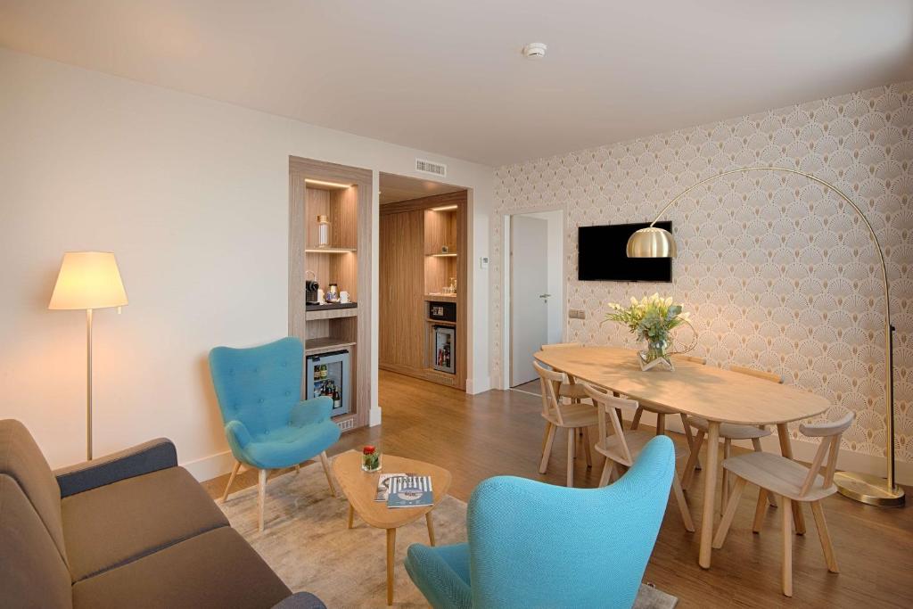 Suite d’hôtel moderne avec un canapé marron, deux fauteuils turquoise et une table basse en bois. Une grande table à manger ovale entourée de chaises assorties se trouve sous un lampadaire arqué, avec une télévision murale au-dessus. Des niches en bois éclairées présentent des objets décoratifs et un minibar. Le papier peint beige à motifs et le parquet créent une ambiance chaleureuse et élégante.