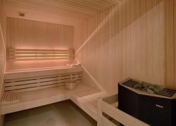 Sauna présentant un intérieur compact entièrement habillé de panneaux en bois naturel clair recouvrant les murs, le plafond et les banquettes intégrées. Deux niveaux d’assises en bois s’étendent le long du fond et du côté gauche, offrant un espace confortable dédié à la détente. Un petit seau en bois accompagné d’une louche est posé sur l’assise inférieure. Au premier plan, à droite, un poêle électrique noir rempli de pierres est protégé par une barrière de sécurité en bois clair. Le sol est recouvert de grands carreaux carrés gris. L’éclairage chaleureux met en valeur les nuances miellées du bois et crée une atmosphère cosy et paisible, propice au bien-être et à la relaxation.