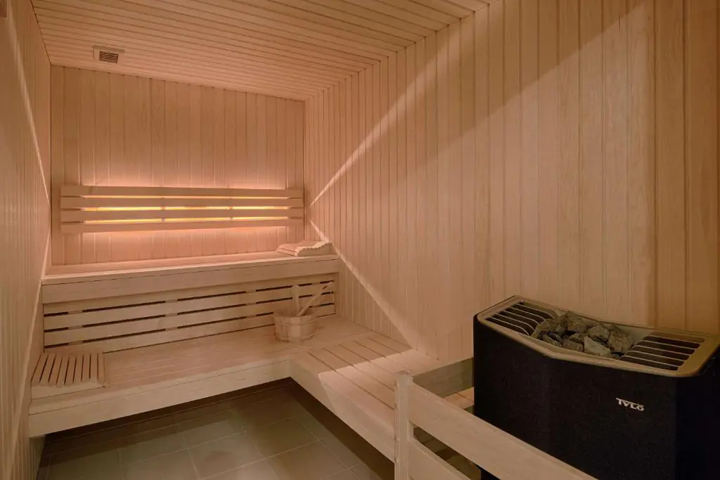Sauna présentant un intérieur compact entièrement habillé de panneaux en bois naturel clair recouvrant les murs, le plafond et les banquettes intégrées. Deux niveaux d’assises en bois s’étendent le long du fond et du côté gauche, offrant un espace confortable dédié à la détente. Un petit seau en bois accompagné d’une louche est posé sur l’assise inférieure. Au premier plan, à droite, un poêle électrique noir rempli de pierres est protégé par une barrière de sécurité en bois clair. Le sol est recouvert de grands carreaux carrés gris. L’éclairage chaleureux met en valeur les nuances miellées du bois et crée une atmosphère cosy et paisible, propice au bien-être et à la relaxation.