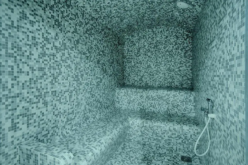 Hammam d’hôtel aménagé dans une pièce entièrement recouverte de petits carreaux de mosaïque carrés aux nuances de turquoise, de bleu pâle, de gris et de vert foncé. Les murs, le plafond voûté, le sol et les banquettes intégrées sont décorés d’un motif géométrique dense en mosaïque. Une longue banquette incurvée longe le mur de gauche et se prolonge vers le fond, tandis que des assises carrelées surélevées offrent un espace de détente. À droite, une douchette à main avec un flexible est fixée au mur carrelé, et une évacuation d’eau est visible au sol. L’éclairage doux aux tonalités froides et l’aménagement fermé créent une atmosphère paisible et intimiste, propice à la relaxation et au bien-être.