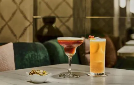 Elegant hotel lounge setting with a sophisticated marble table, featuring two beautifully presented cocktails, one in a refined coupe glass with layered red tones and another in a tall glass with a vibrant golden-orange hue. A small porcelain bowl of assorted nuts accompanies the drinks, while plush green velvet seating and warm decorative details create an intimate, upscale atmosphere.