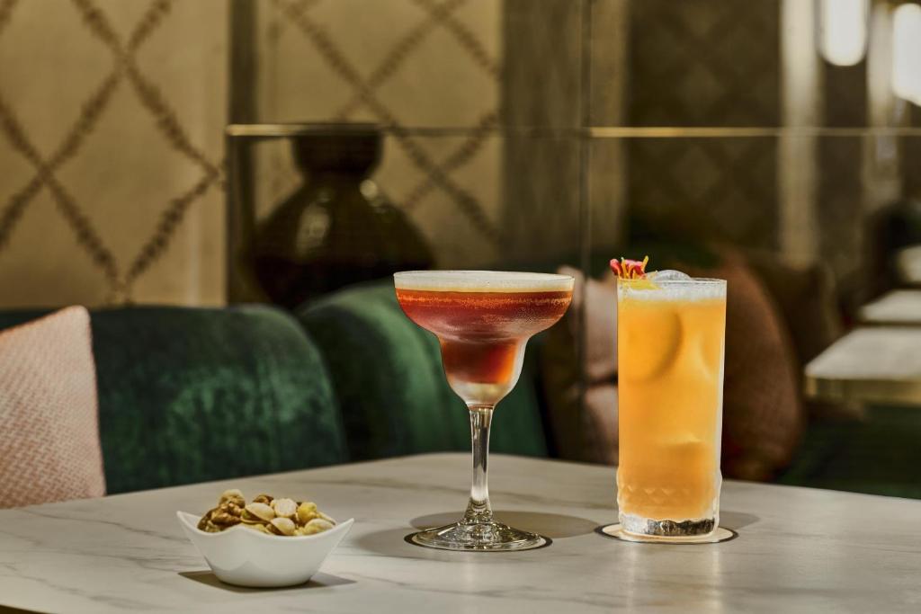 Elegant hotel lounge setting with a sophisticated marble table, featuring two beautifully presented cocktails, one in a refined coupe glass with layered red tones and another in a tall glass with a vibrant golden-orange hue. A small porcelain bowl of assorted nuts accompanies the drinks, while plush green velvet seating and warm decorative details create an intimate, upscale atmosphere.