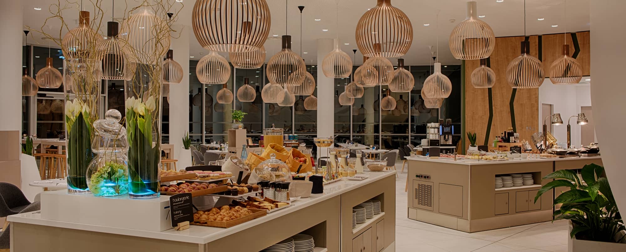 Salle de petit-déjeuner d’hôtel composée de deux grands buffets en marbre blanc présentant des viennoiseries, du pain, des fromages et différents jus de fruits. Des tulipes blanches et des plantes vertes disposées dans des vases lumineux décorent les comptoirs. De nombreux luminaires modernes aux finitions effet bois surplombent la salle. Au fond, plusieurs tables rondes sont installées près de grandes baies vitrées. Les murs latéraux présentent un revêtement effet bois, tandis que les murs et les colonnes sont de couleur crème. Le sol est recouvert de grands carreaux de carrelage blanc.