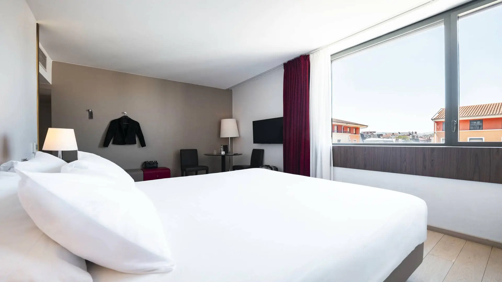 A hotel room with views over the city rooftops, burgundy curtains, and a black jacket hanging from a wall hook.