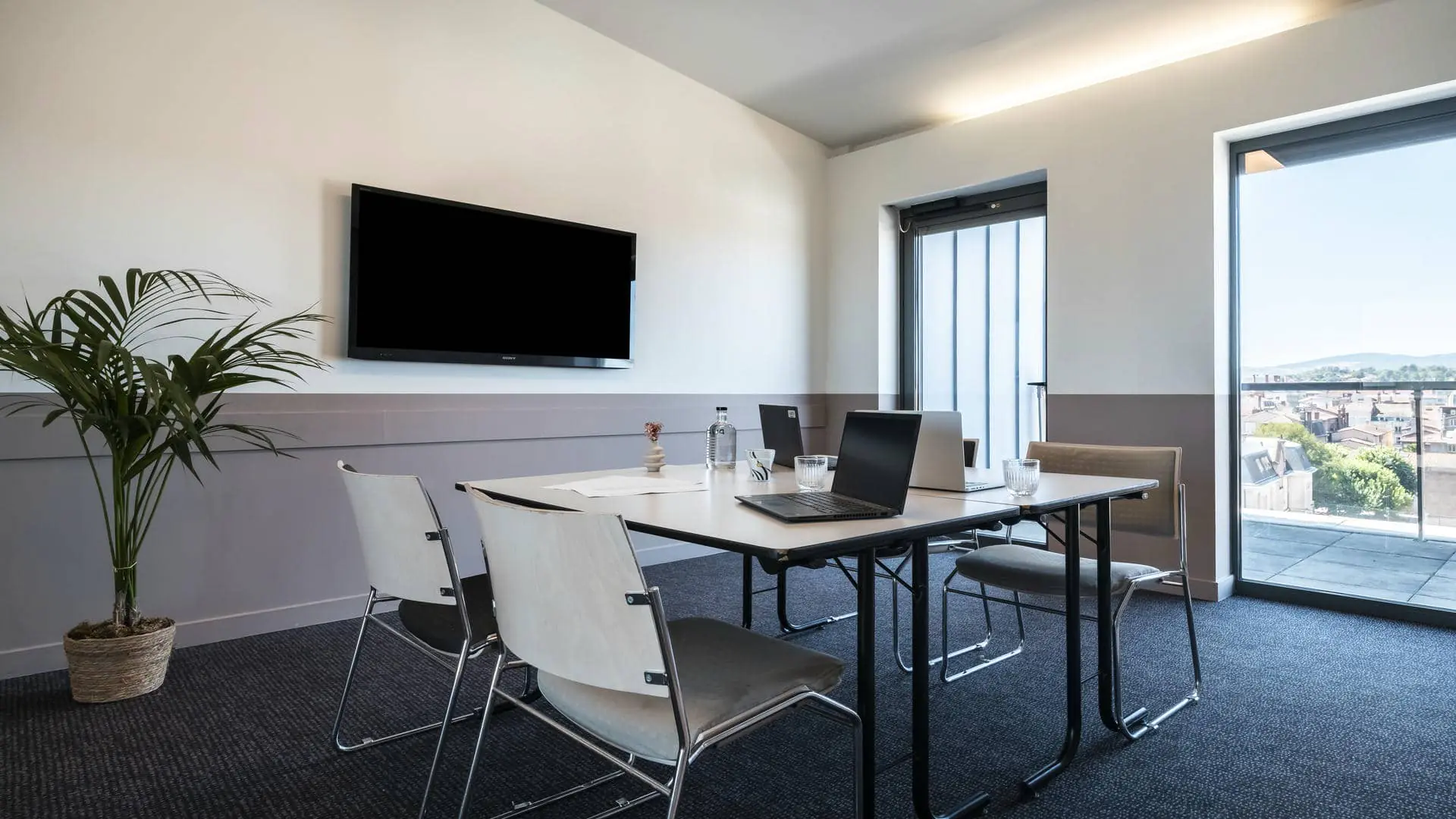 Meeting room equipped with a black wall-mounted television screen, two white work tables, four black-and-white fabric chairs, a dedicated break area, and two large glass doors overlooking the hotel’s rooftop terrace.