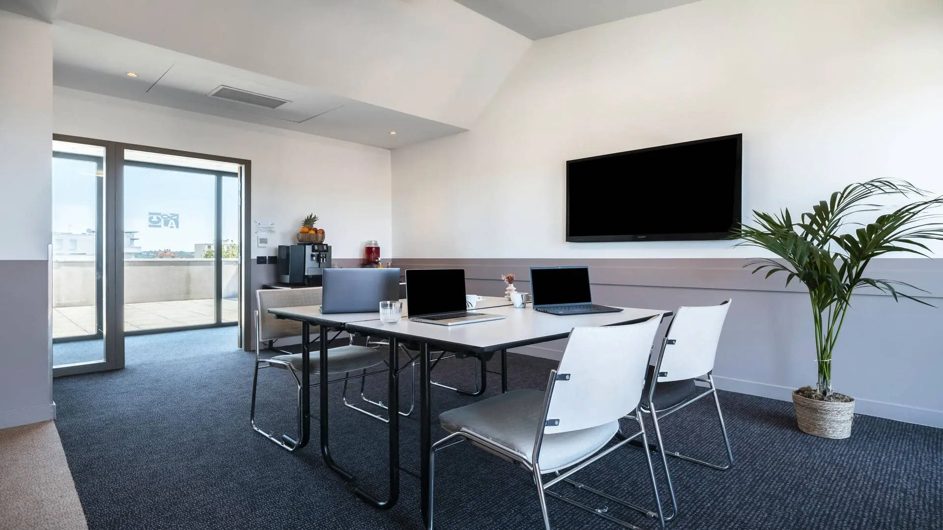 Training room equipped with a black wall-mounted television screen, two white work tables, four black-and-white fabric chairs, a dedicated break area, and open French doors leading to the corridor and providing direct access to the rooftop terrace.
