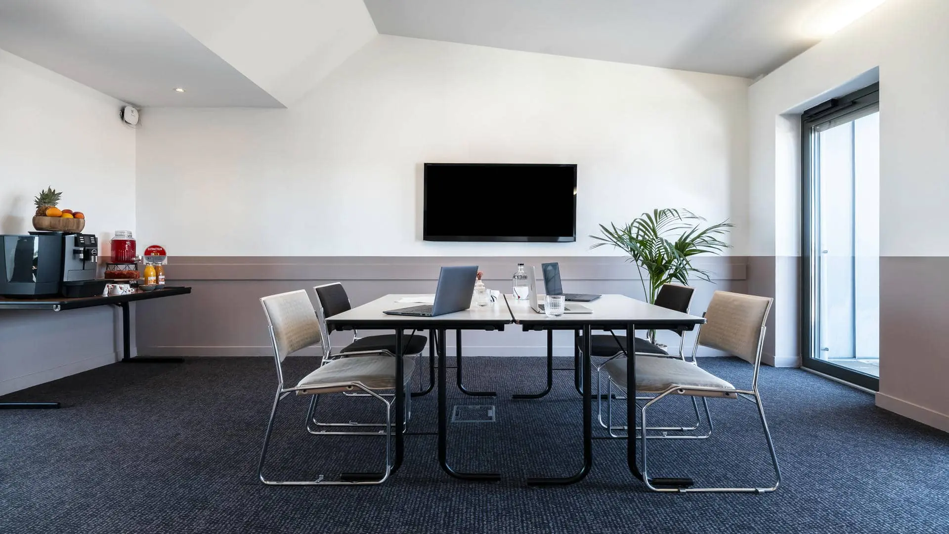Training room equipped with a black wall-mounted television screen, two white work tables, four black-and-white fabric chairs, a dedicated break area, and direct access to the rooftop terrace.