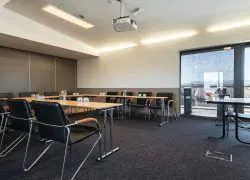 Meeting room with large floor-to-ceiling windows fitted with blinds, offering views over the city rooftops, wooden tables arranged in a U-shape, and black leather chairs positioned around them.