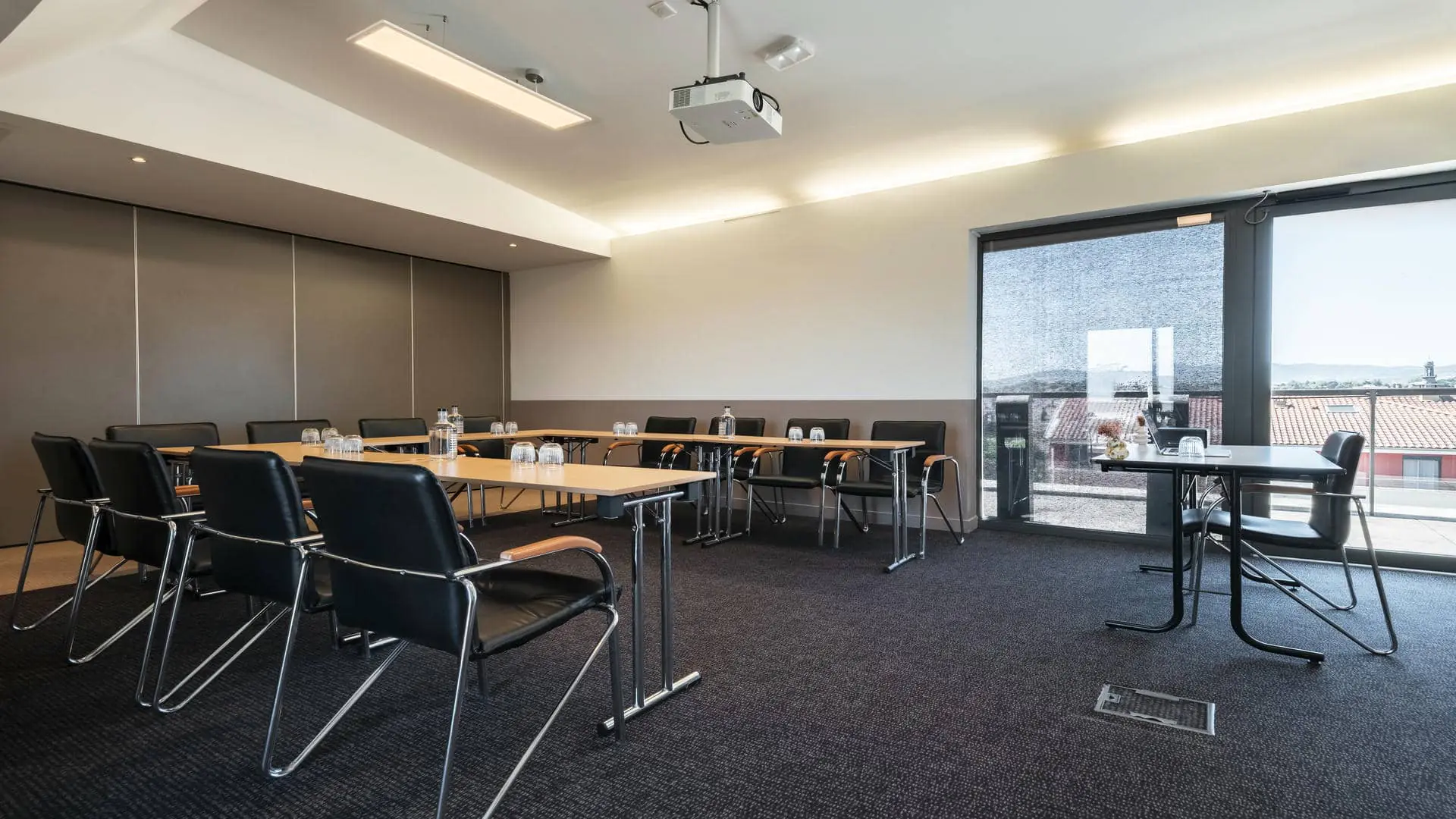 Meeting room with large floor-to-ceiling windows fitted with blinds, offering views over the city rooftops, wooden tables arranged in a U-shape, and black leather chairs positioned around them.