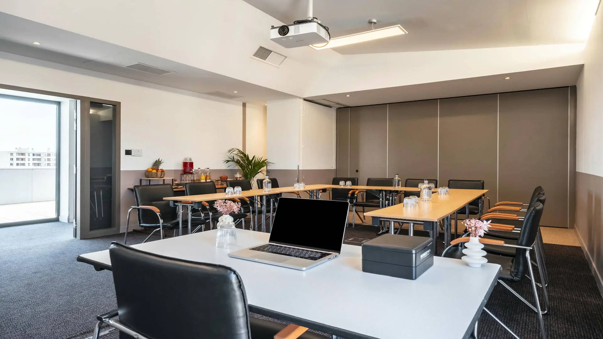 Meeting room rental with carpeted flooring, white walls up to mid-height and beige above, tables arranged in a U-shape, and open doors providing direct access to the rooftop with views over the city and the Beaujolais region.