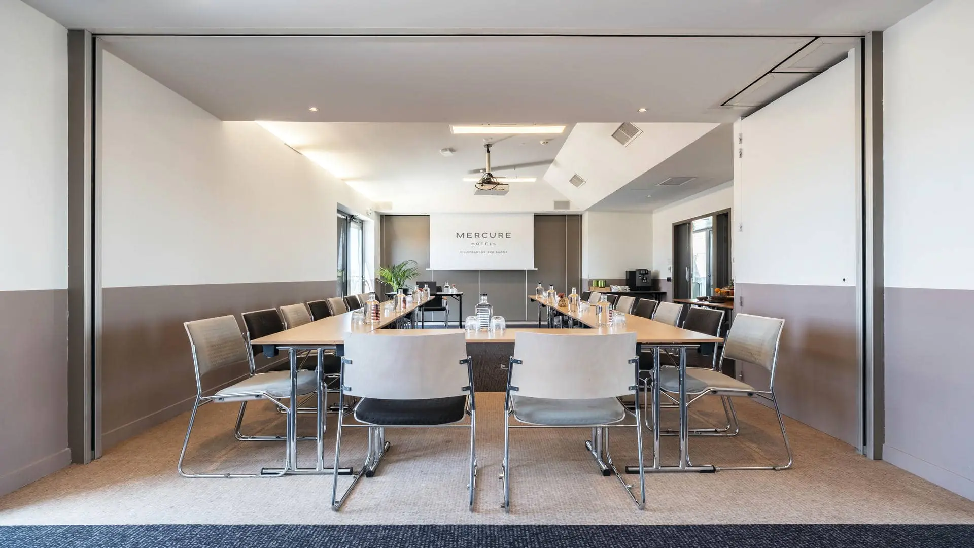 Seminar room rental with carpeted flooring, white walls up to mid-height and beige above, a projection screen featuring the Mercure Villefranche-sur-Saône logo, tables arranged in a U-shape, and direct access to the rooftop.