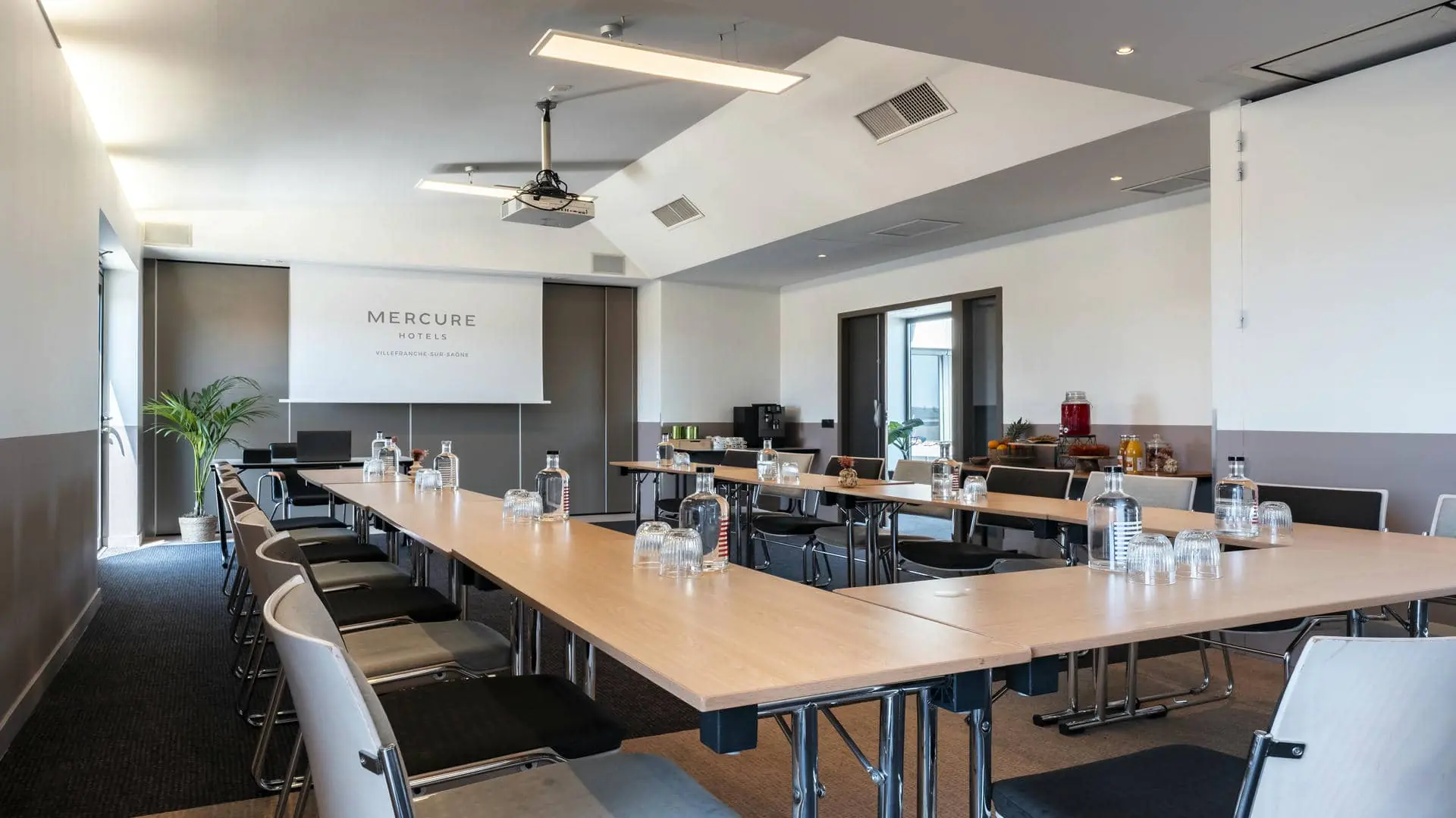 Seminar room rental with carpeted flooring, white walls up to mid-height and beige above, a projection screen featuring the Mercure Villefranche-sur-Saône logo, tables arranged in a U-shape, and open doors leading to the corridor’s large windows with direct access to the hotel’s rooftop terrace.