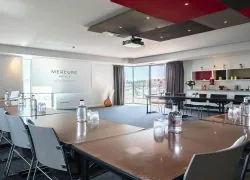 A conference room with tables and chairs arranged in a U-shape, a projection screen featuring the Mercure Villefranche-sur-Saône logo, and views of the hotel’s rooftop terrace.