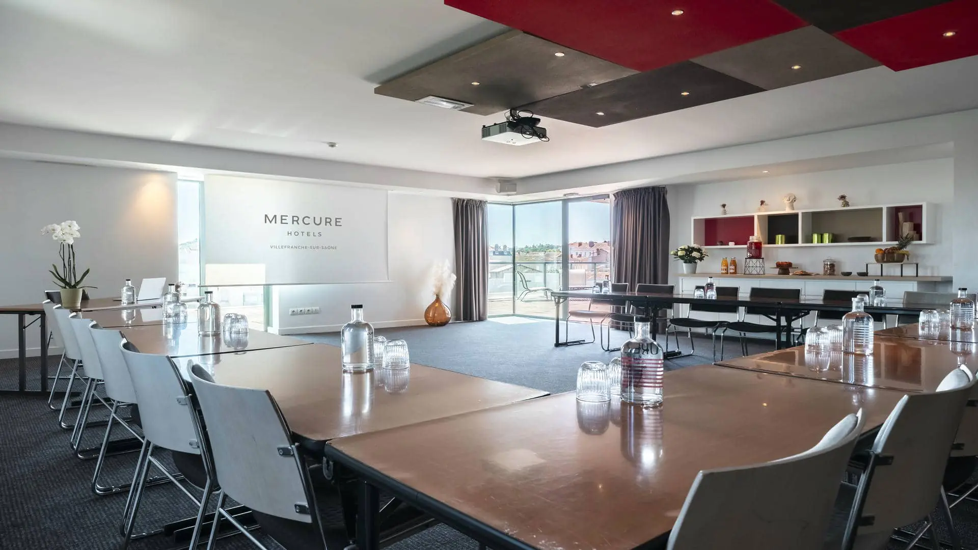 A conference room with tables and chairs arranged in a U-shape, a projection screen featuring the Mercure Villefranche-sur-Saône logo, and views of the hotel’s rooftop terrace.