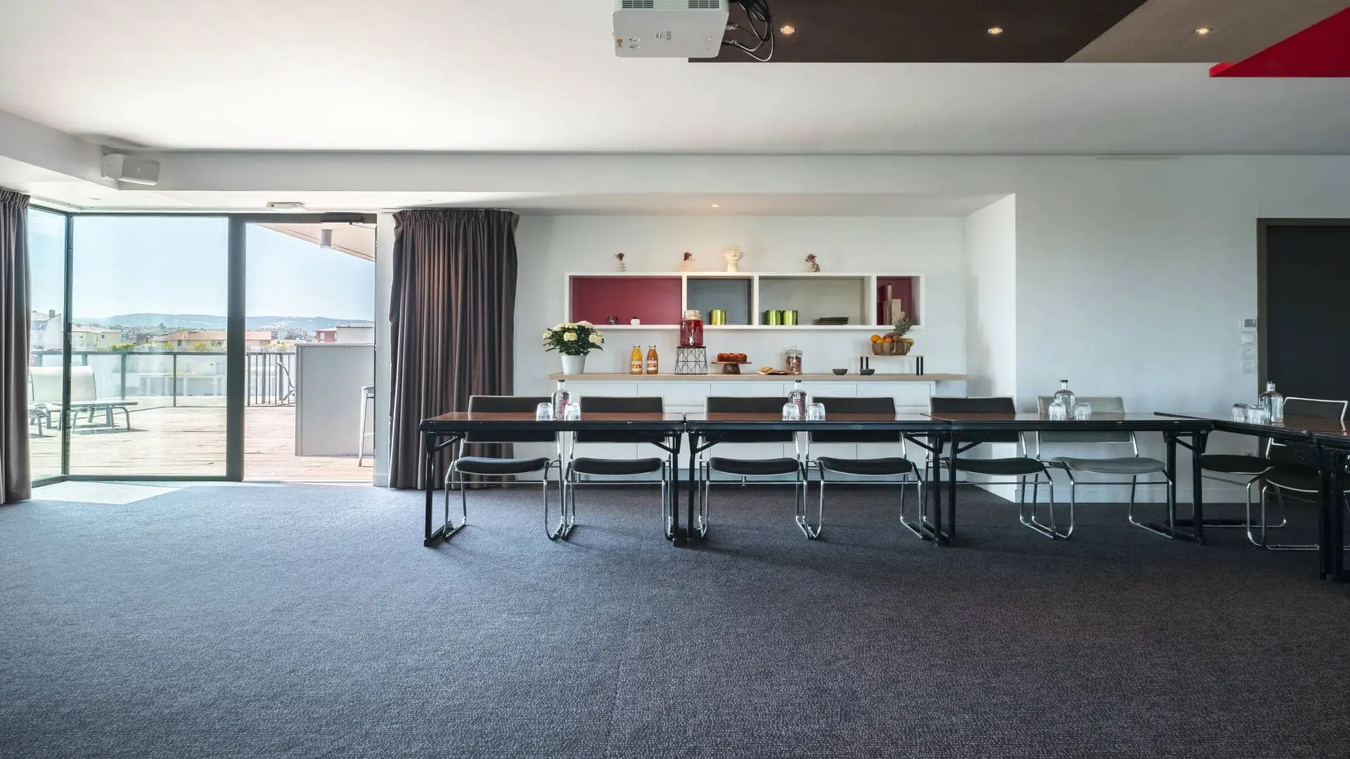A conference room with tables and chairs arranged in a U-shape, a projection screen featuring the Mercure Villefranche-sur-Saône logo, and views of the hotel’s rooftop terrace.