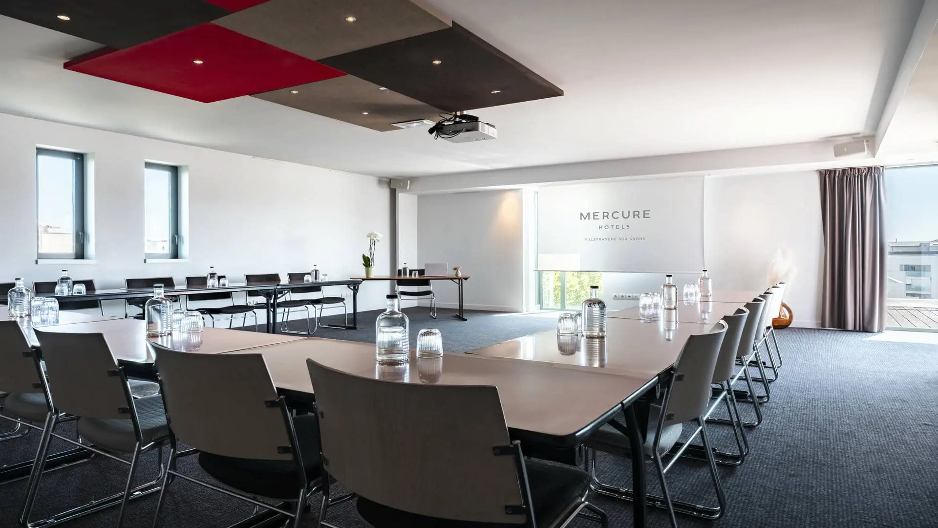 A conference room with tables and chairs arranged in a U-shape, a projection screen featuring the Mercure Villefranche-sur-Saône logo, and views of the hotel’s rooftop terrace.